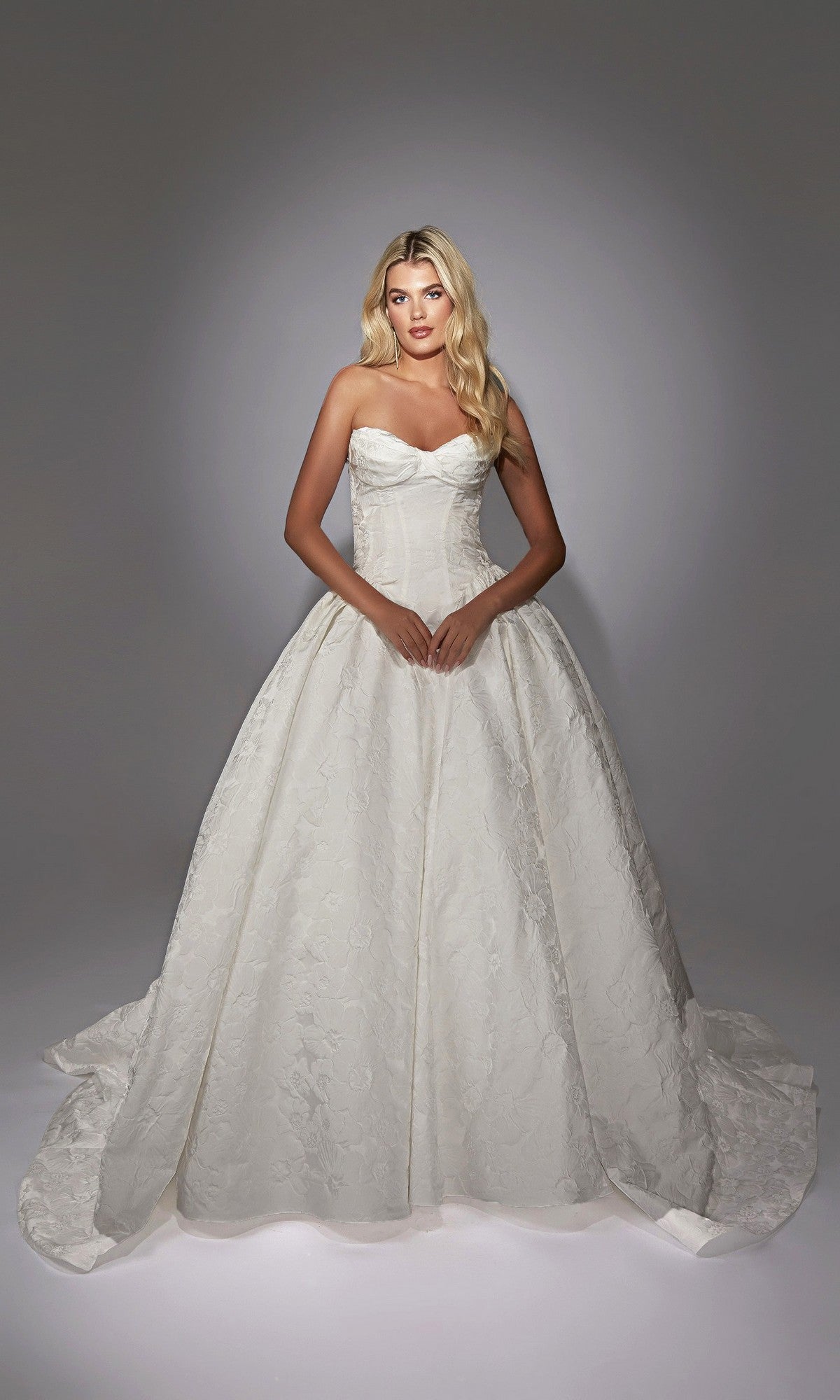 A woman stands against a plain gradient background wearing the Long White Dress: Alyce 7164, a strapless white ball gown with a fitted bodice, full skirt, and elegant floral jacquard fabric for luxurious texture and style.